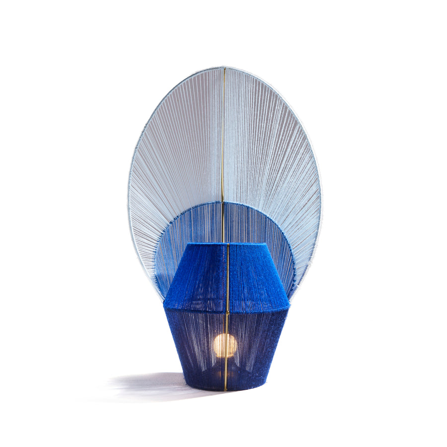 The Ames Farol Side Lamp by Ames is a modern blue table lamp, handmade in Colombia with a geometric base and a large fan-shaped shade wrapped in blue and white string, shown against a white background.