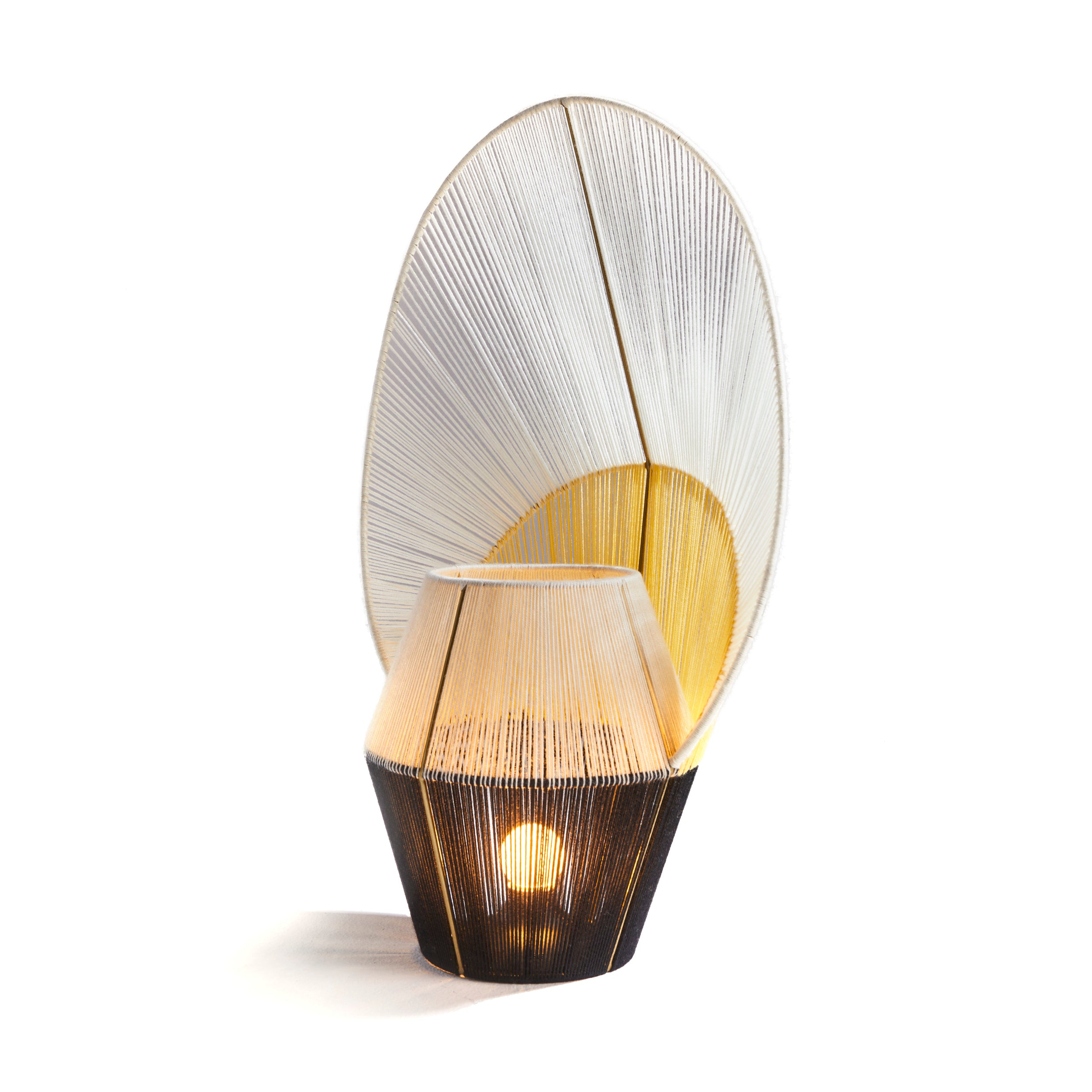 The Ames Farol Side Lamp by Ames is a handmade decorative lamp from Colombia with a geometric base and large fan-shaped woven shade in white and yellow, emitting warm light and showcasing its intricate Farols-inspired design.