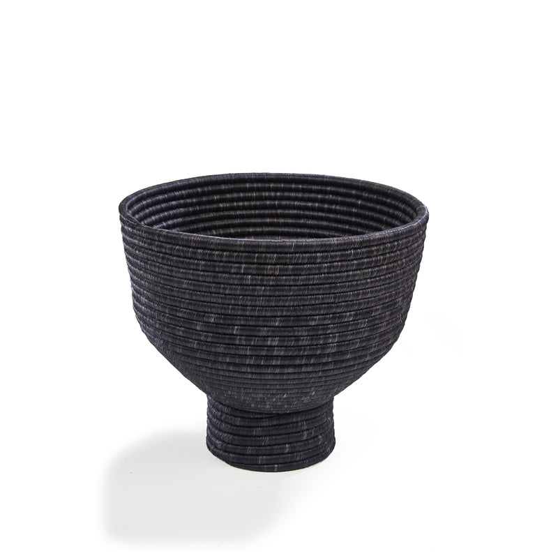 The Ames La Che Pot by Ames is a black, textured bowl with a wide rim and narrow base, inspired by Colombian culture. Horizontal ridges encircle its surface and it stands out against a plain white background.