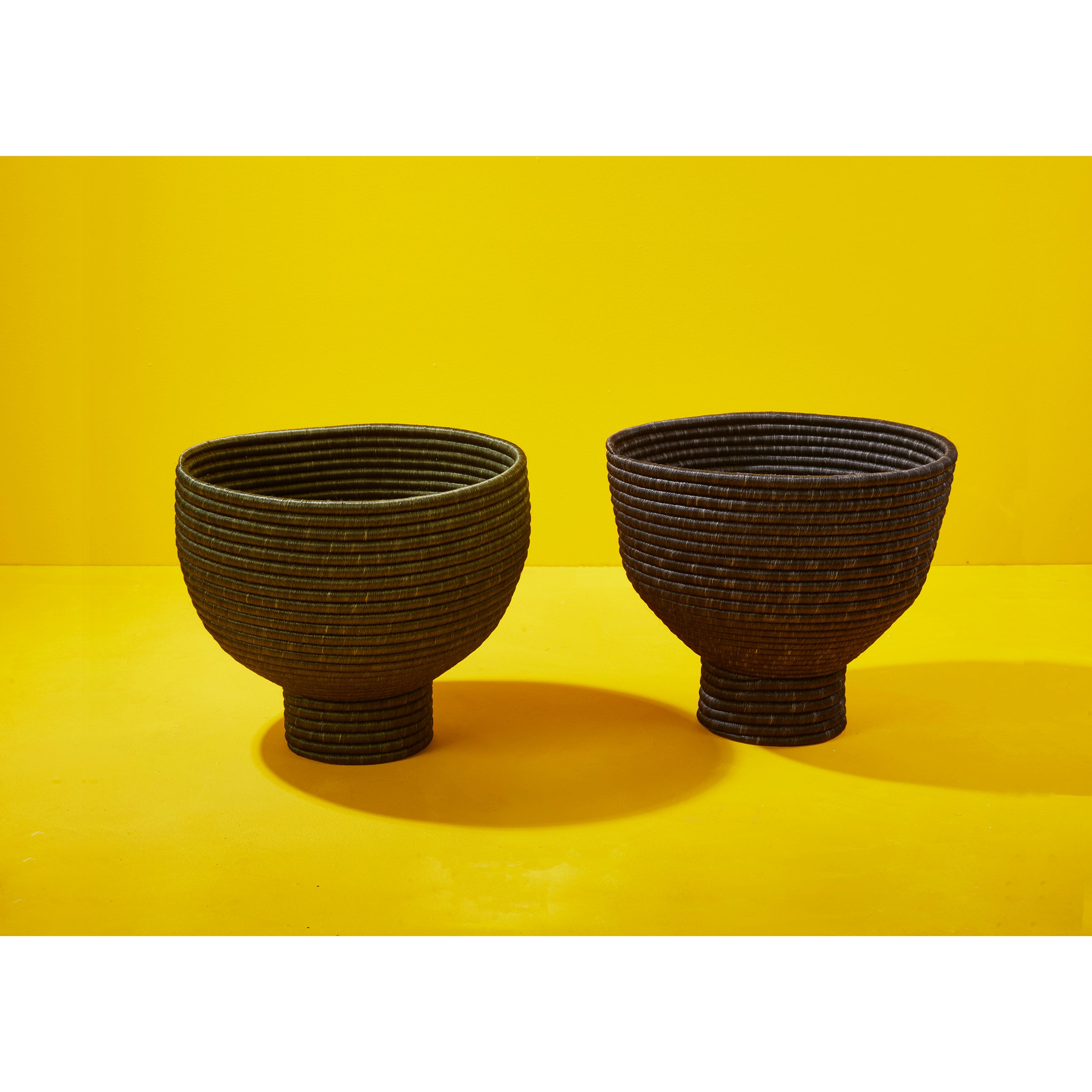 Two dark, textured, round Ames La Che Pots by Ames, crafted from fique fiber, sit side by side on a bright yellow surface with a matching background, showcasing Colombian artistry.