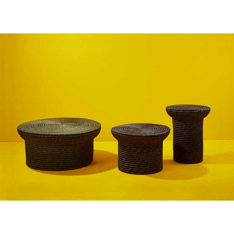 Against a bright yellow background, three textured, round tables of varying heights and sizes are showcased. The La Che sidetable by Ames, designed by Sebastian Herkner, exhibits a dark ribbed pattern reminiscent of traditional weaving craft, contrasting strikingly with the backdrop.