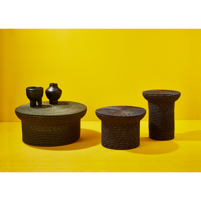 Three textured, dark-brown round tables by Ames vary in height against a vibrant yellow background. The left table features two small black ceramic vases. Designed by Sebastian Herkner, the La Che sidetable showcases a circular, traditional weaving craft pattern.