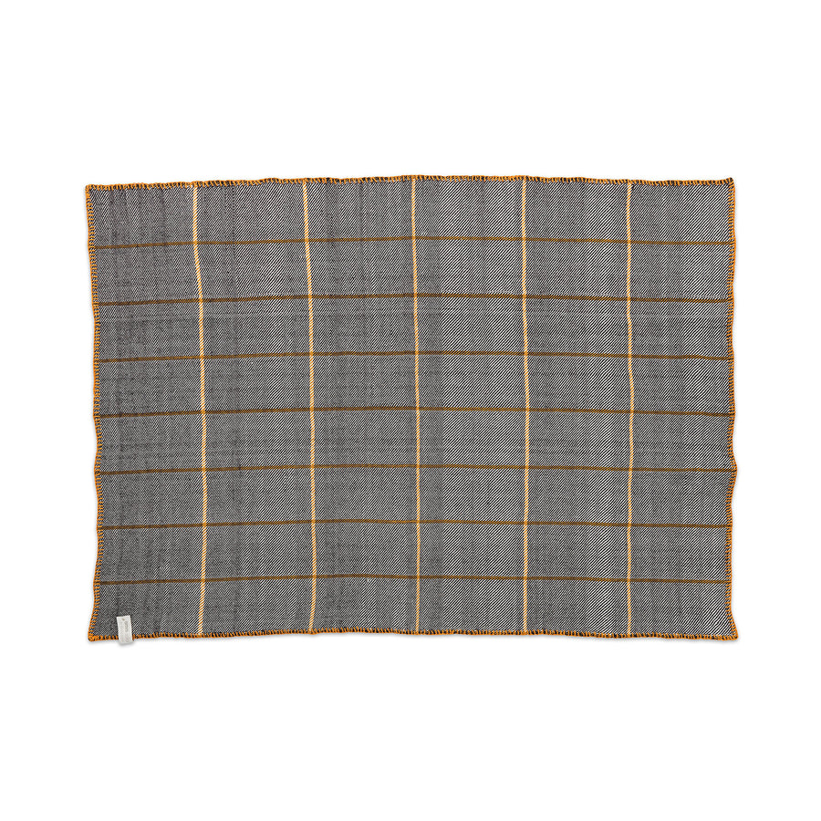 The Ames Manta Plaid by Ames is a rectangular gray textile with a grid of thin yellow lines and yellow stitching, laid flat on white. Showcasing Colombian craftsmanship, it features a small fabric tag on one corner.