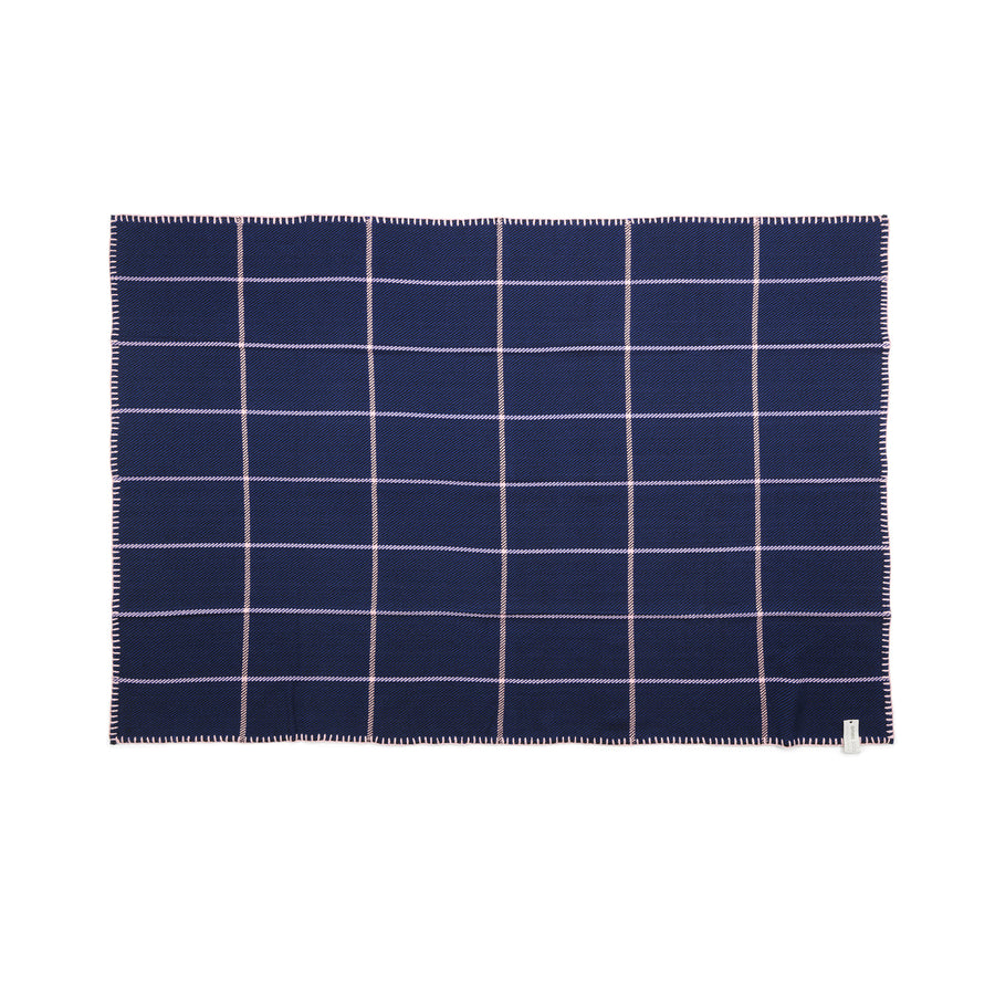 The Ames Manta Plaid by Ames is a rectangular navy blue blanket with a white grid pattern, whipstitched edges, and a small label at the lower right corner. Inspired by luxury wool throws, it's shown flat against a white background.