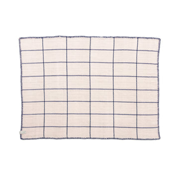 The Ames Manta Plaid by Ames is a square cream-colored cloth napkin featuring a navy blue grid pattern and stitched edges, reflecting traditional Colombian craftsmanship, shown flat on a white background.