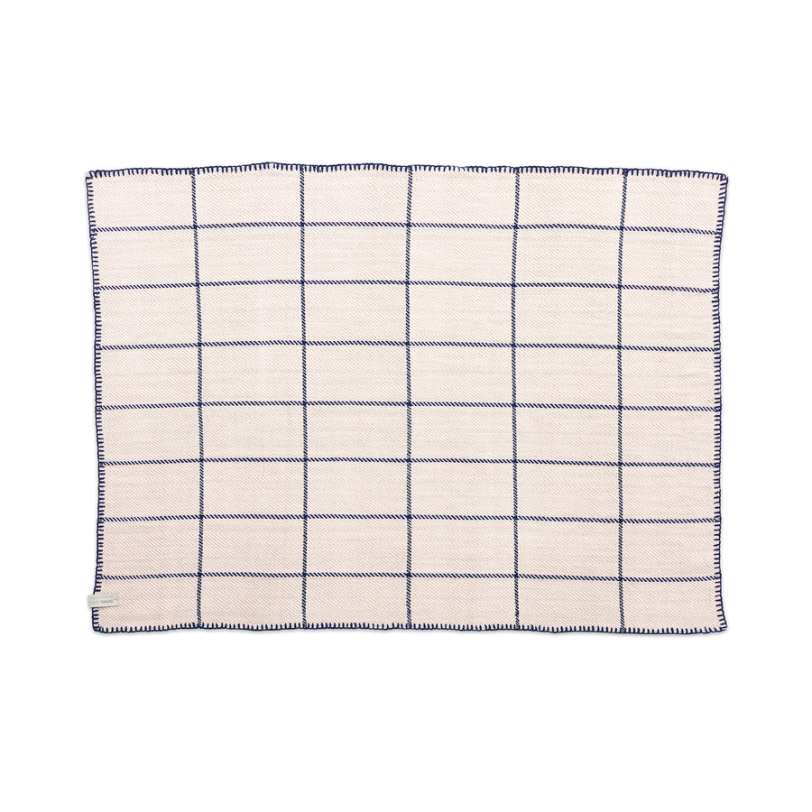 The Ames Manta Plaid by Ames is a square cream-colored cloth napkin featuring a navy blue grid pattern and stitched edges, reflecting traditional Colombian craftsmanship, shown flat on a white background.