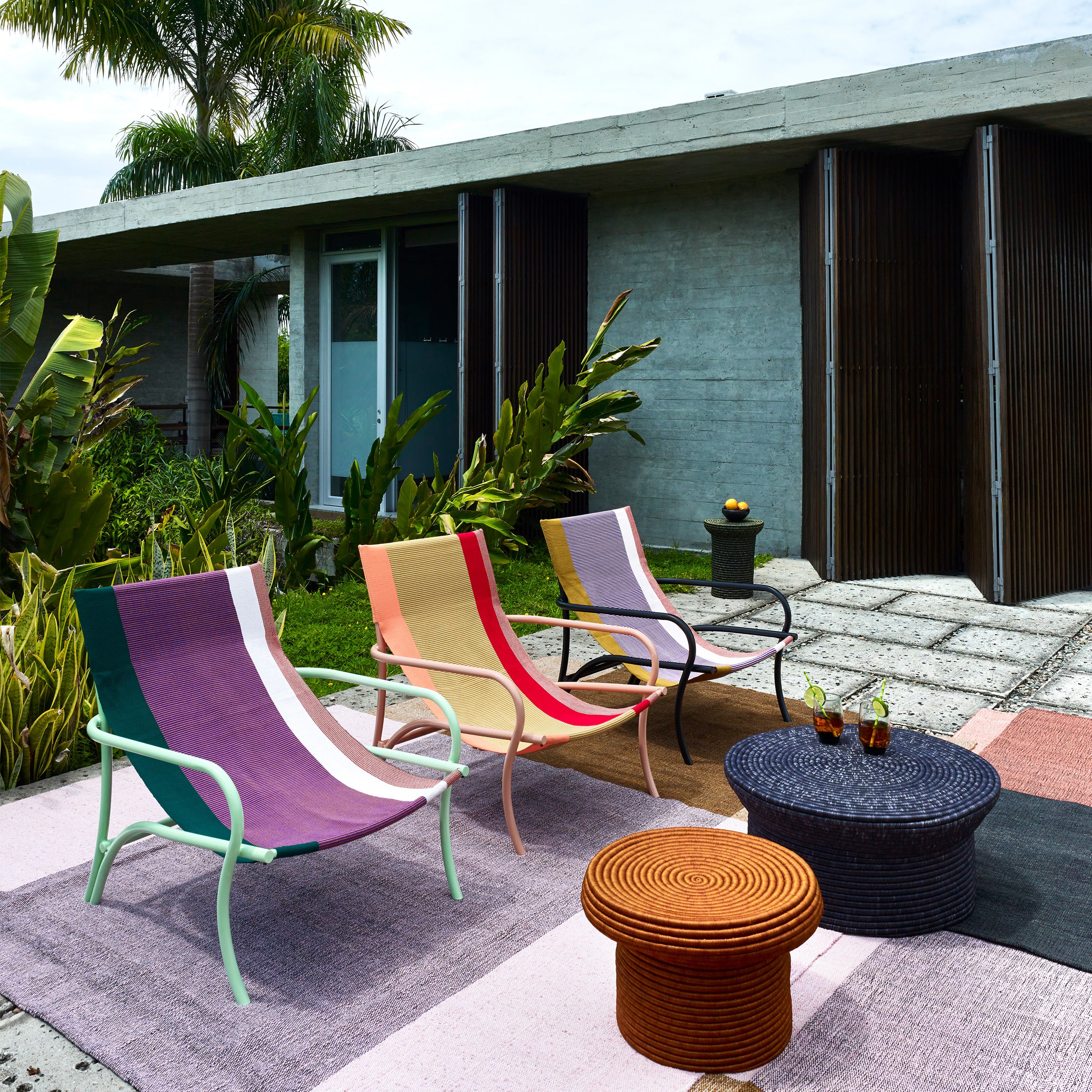 A modern outdoor patio showcases three Ames Maraca Lounge Chairs, vibrant on an ethnic-patterned rug. A round wicker coffee table holds drinks and a fruit bowl, surrounded by lush greenery with a concrete building as the backdrop.