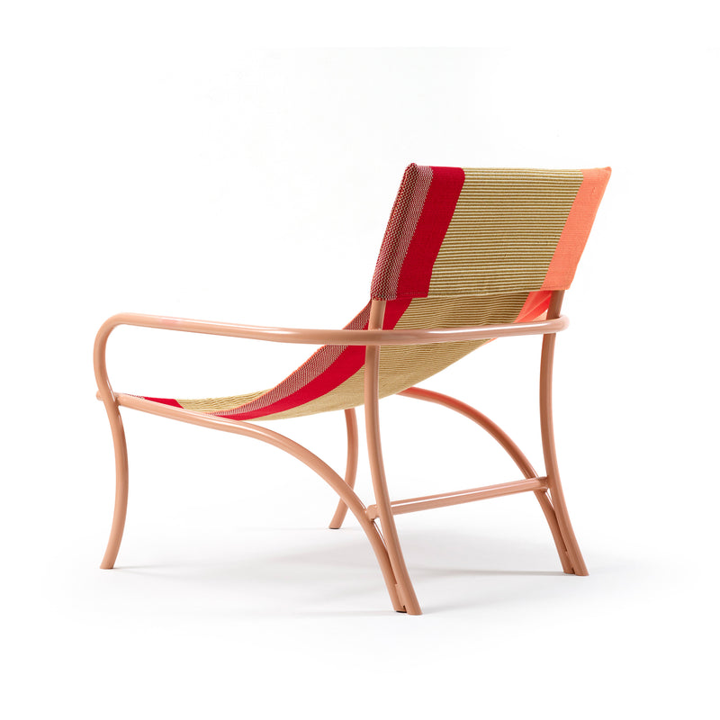 The Maraca Lounge Chair by Ames features a modern design with a light pink metal frame, and a woven seat and backrest in beige, red, and orange stripes. Its minimalist, gently curved style is reminiscent of Colombian hammocks, set against a plain white background.