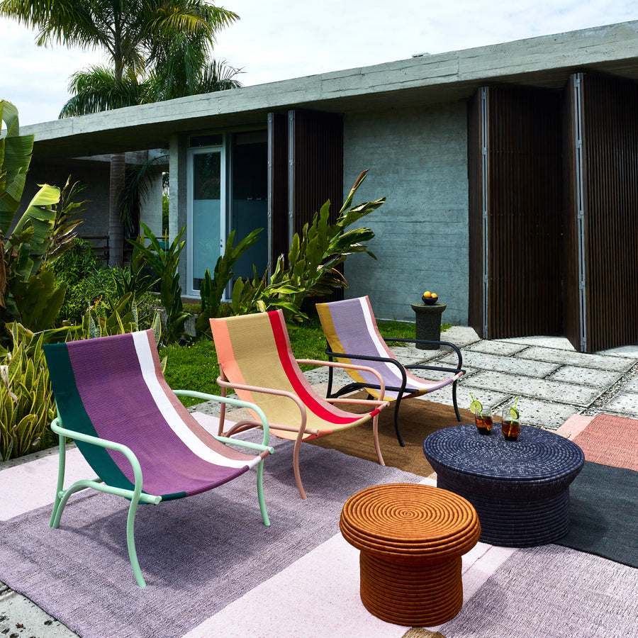 A modern outdoor patio showcases three Ames Maraca Lounge Chairs, vibrant on an ethnic-patterned rug. A round wicker coffee table holds drinks and a fruit bowl, surrounded by lush greenery with a concrete building as the backdrop.
