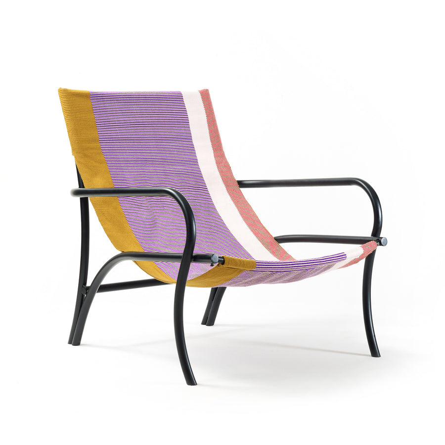 The Maraca Lounge Chair by Ames features a black metal frame and a vibrant fabric seat, reminiscent of Colombian hammocks, with stripes of purple, mustard yellow, white, and pink. Its seamless curved backrest and seat are set against a plain white background.
