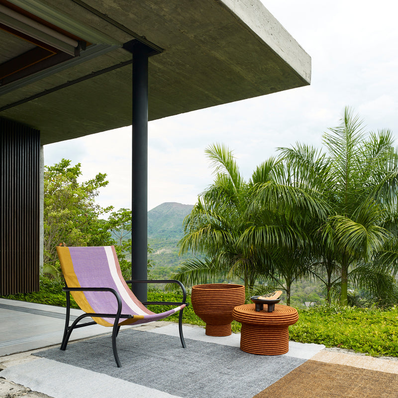 A contemporary patio showcases the vibrant Maraca Lounge Chair by Ames, paired with two wicker stools on a rug. The space, with lush greenery and mountain views, is enclosed by a concrete roof and glass door. Tall palm trees sway under the cloudy sky, echoing subtle ethnic designs.