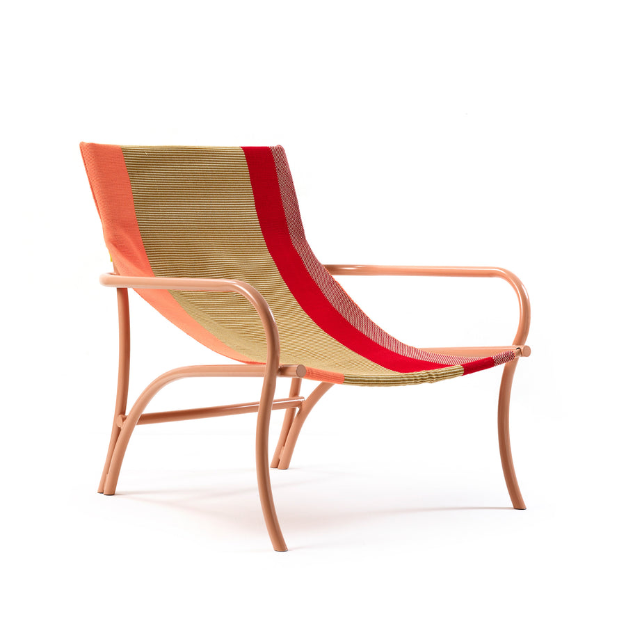 The Maraca Lounge Chair by Ames features a minimalist metal frame in light pink, inspired by Colombian hammocks. Its sleek, curved design has a woven fabric seat and backrest with earthy tones of orange, beige, and bold red stripes for an ethnic touch.