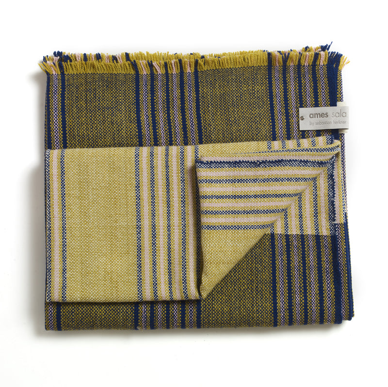 The Ames Mulera Throw by Ames features olive green, yellow, and blue stripes with fringed edges. Neatly folded, it displays both its striped front and solid back, with a corner label that reads "ames sala by sebastian herkner.
