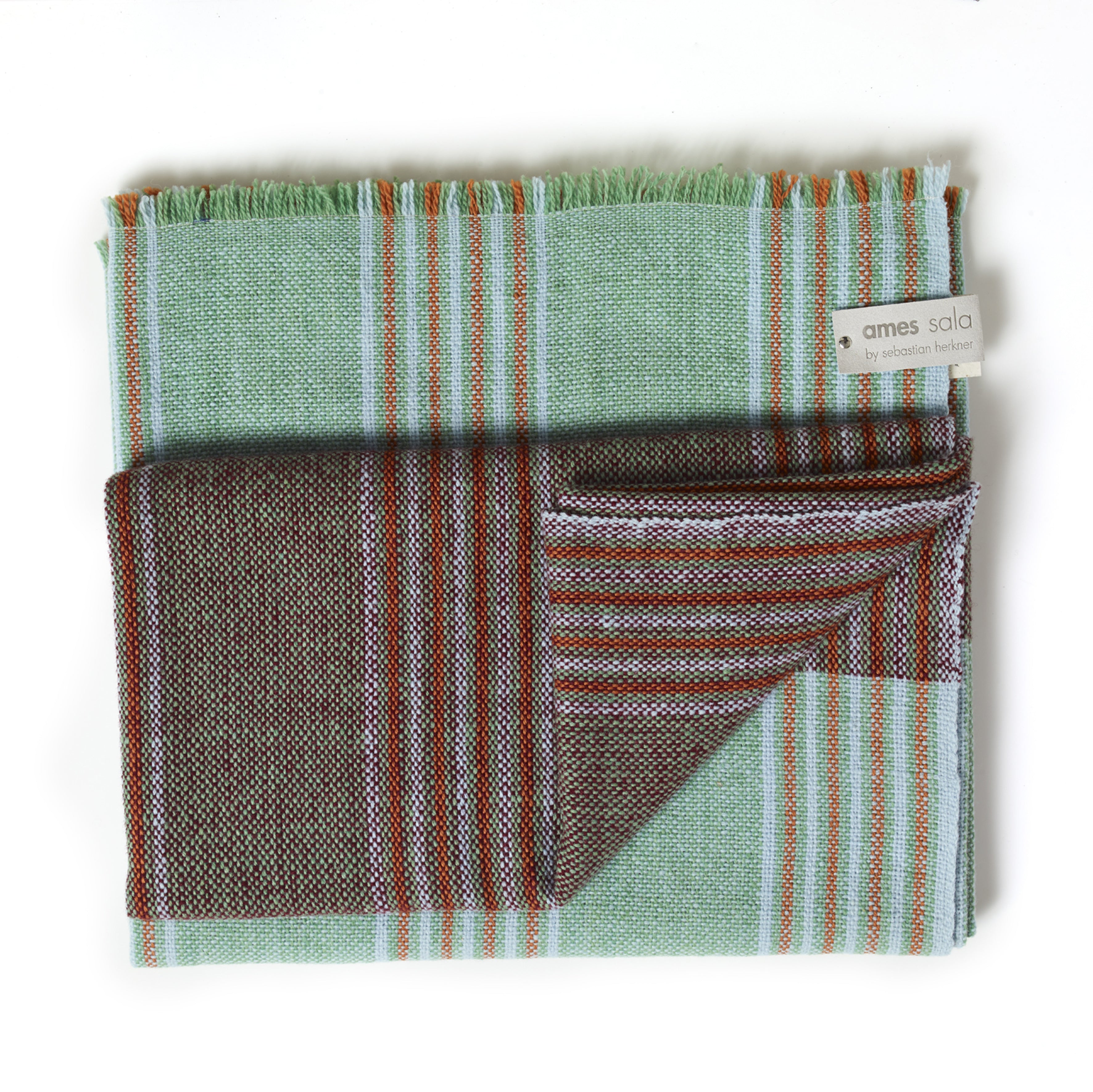 A folded Ames Mulera Throw by Ames, featuring green, brown, blue, and red stripes, fringed edges, and a visible "ames sala by sebastian herkner" fabric label.