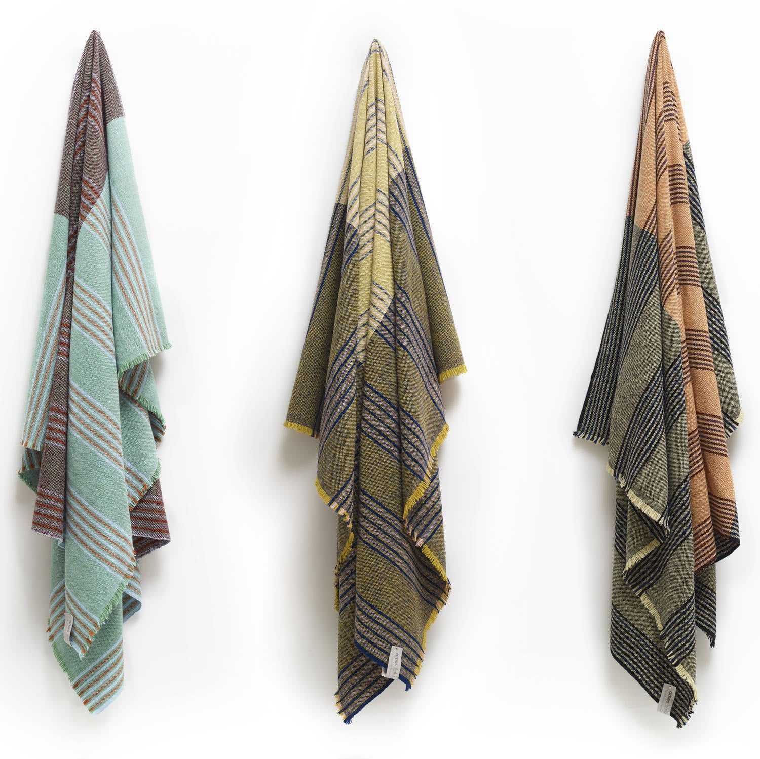 Three Ames Mulera Throws by Ames in pastel green, yellow, and tan hang side by side on a white wall, each lightweight with fringed edges and draped in a triangular shape.