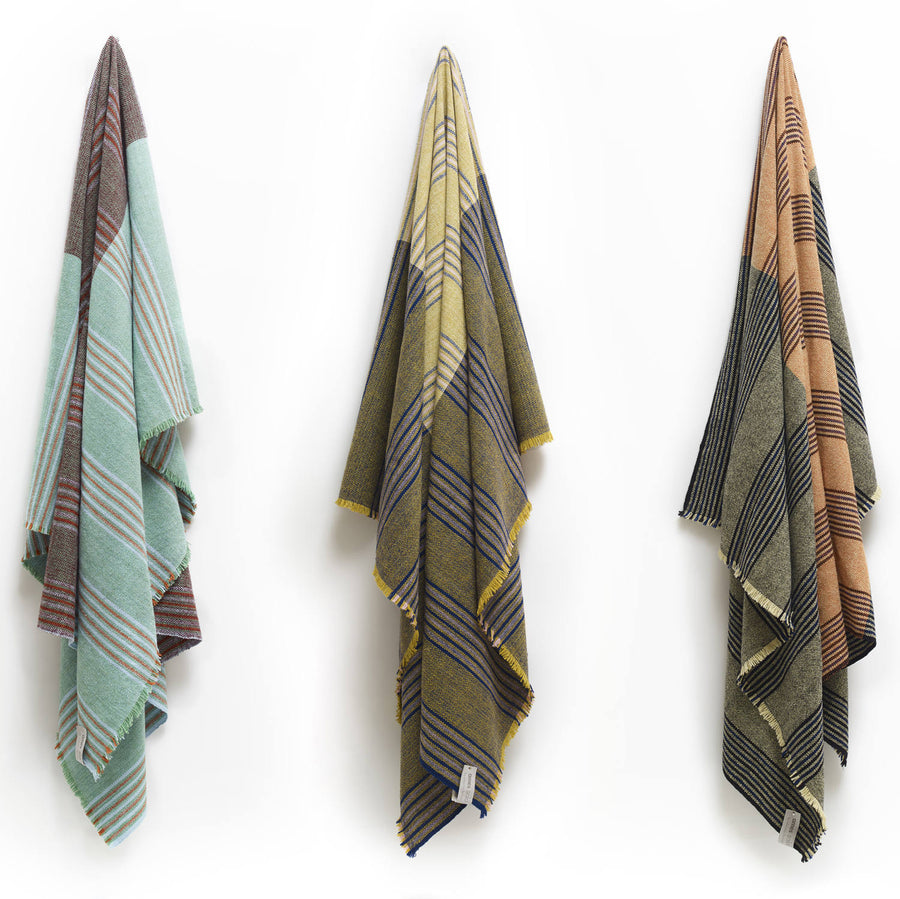 Three Ames Mulera Throws by Ames in pastel green, yellow, and tan hang side by side on a white wall, each lightweight with fringed edges and draped in a triangular shape.