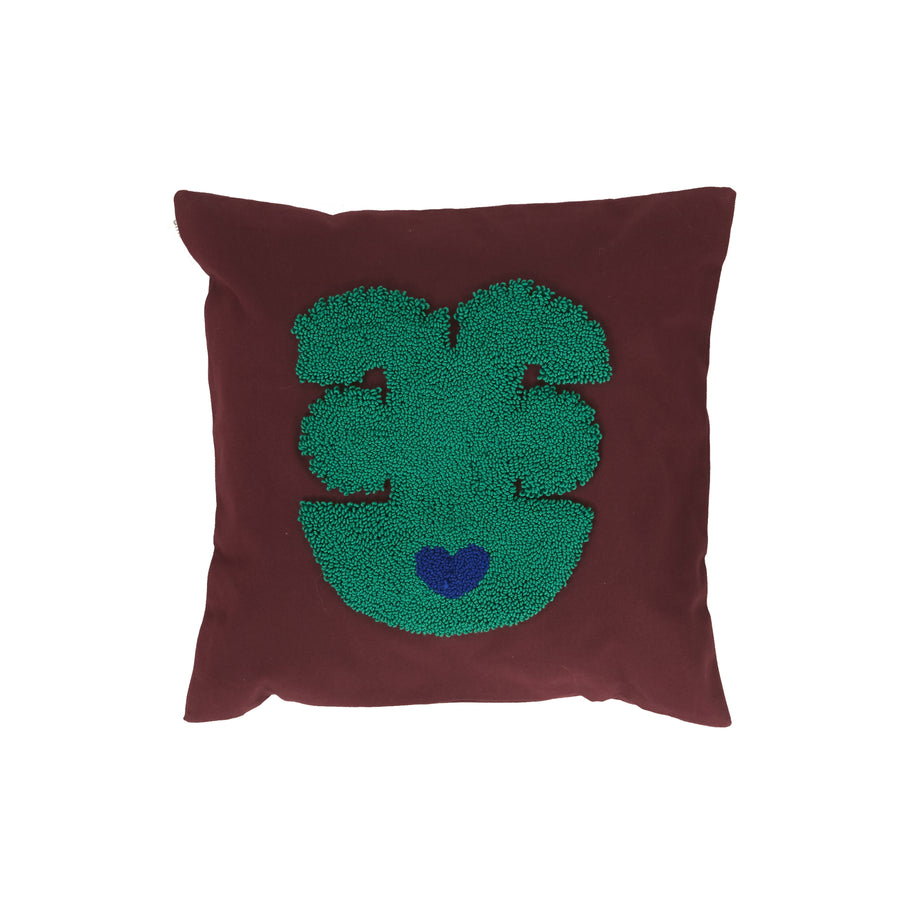 Part of Ames Nido Qeztal collection, this square cushion features a brown cover with a textured abstract green face design centered around a blue heart-shaped nose on a plain white background.