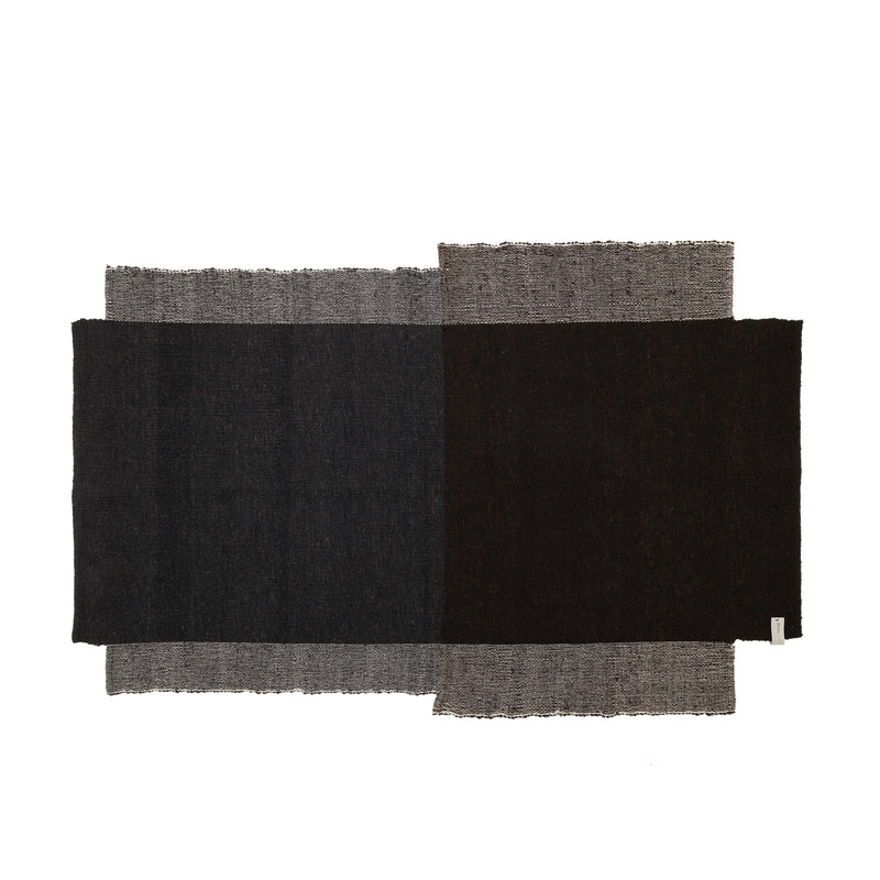 The Nobsa Rug by Ames is a hand-woven, rectangular wool textile. It features an abstract design with overlapping dark gray, black, and light brown sections, creating a textured look with frayed edges that reflect the artisanal heritage of Colombias city of Nobsa.