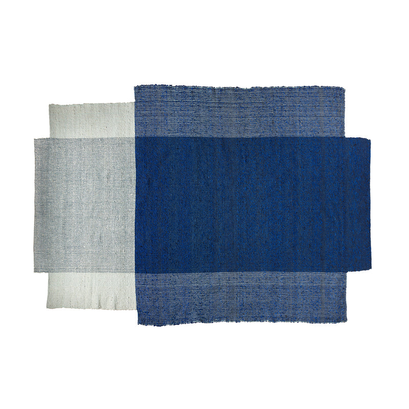 The Nobsa Rug by Ames is a textile art piece with three overlapping wool panels: deep blue, medium blue, and light gray. Inspired by Nobsa rugs from the Colombian city of Nobsa, it forms a layered geometric composition.