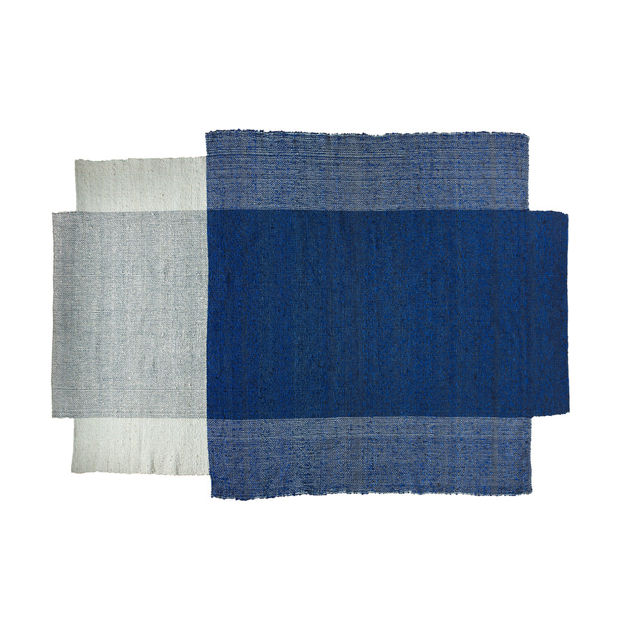 The Nobsa Rug by Ames is a textile art piece with three overlapping wool panels: deep blue, medium blue, and light gray. Inspired by Nobsa rugs from the Colombian city of Nobsa, it forms a layered geometric composition.