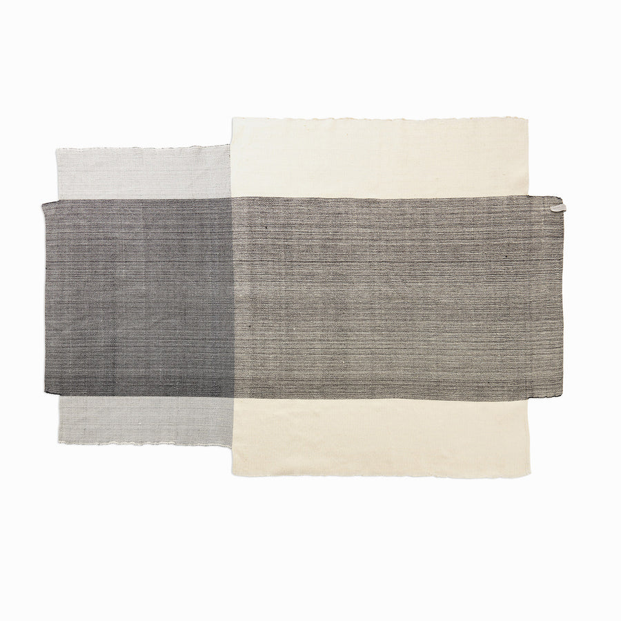 The Nobsa Rug by Ames is a minimalist textile featuring overlapping rectangles in gray, black, and beige, echoing the layered grid patterns of traditional rugs from Nobsa, Colombia. This exquisite piece is hand-woven from fine wool on a white background.