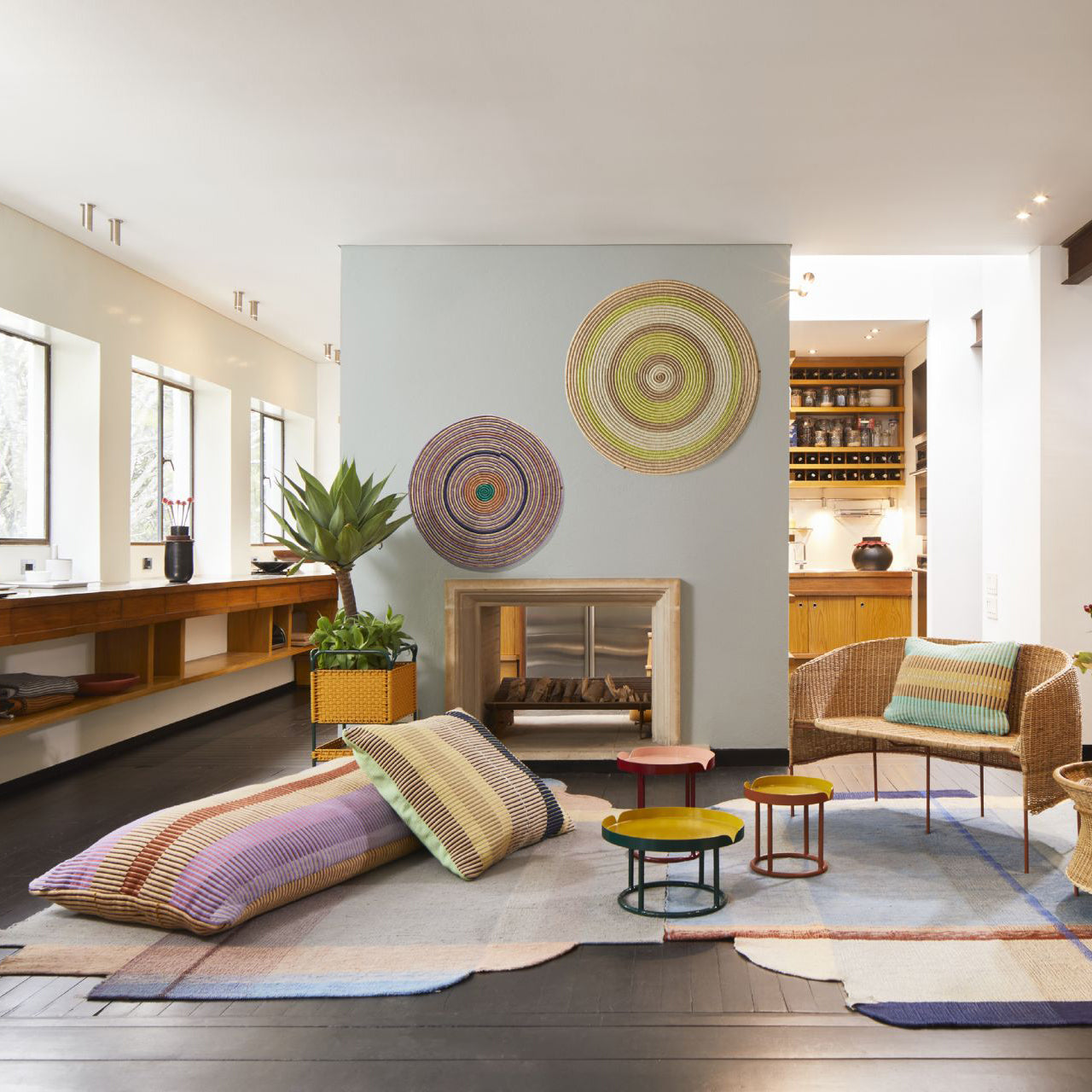 A modern living room with colorful woven rugs, low cushions, small round tables, and a wicker chair with a striped pillow. Above the fireplace, Ames Ojitos Wall Decoration Andes by Ames adds artistic flair and warmth to the naturally lit space.