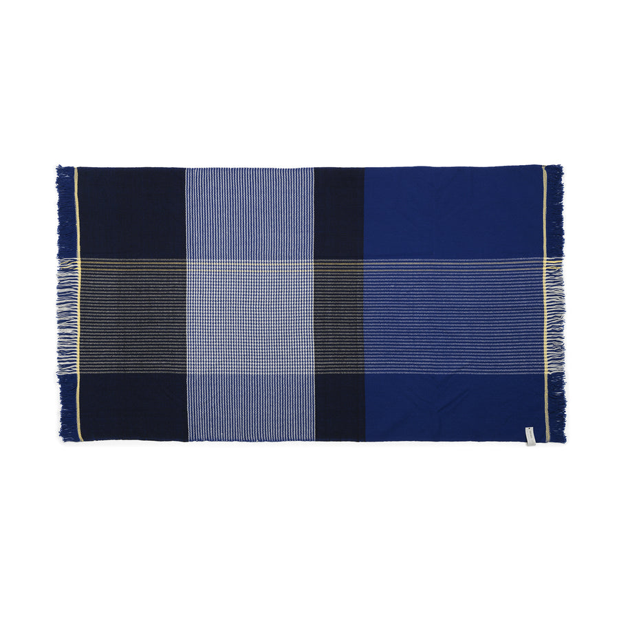 The Ames Ruana Throw, featuring traditional craft techniques, combines solid, striped, and checkered patterns in blue and black. Fringed edges enhance its classic charm and a small tag on one side completes the elegant look.