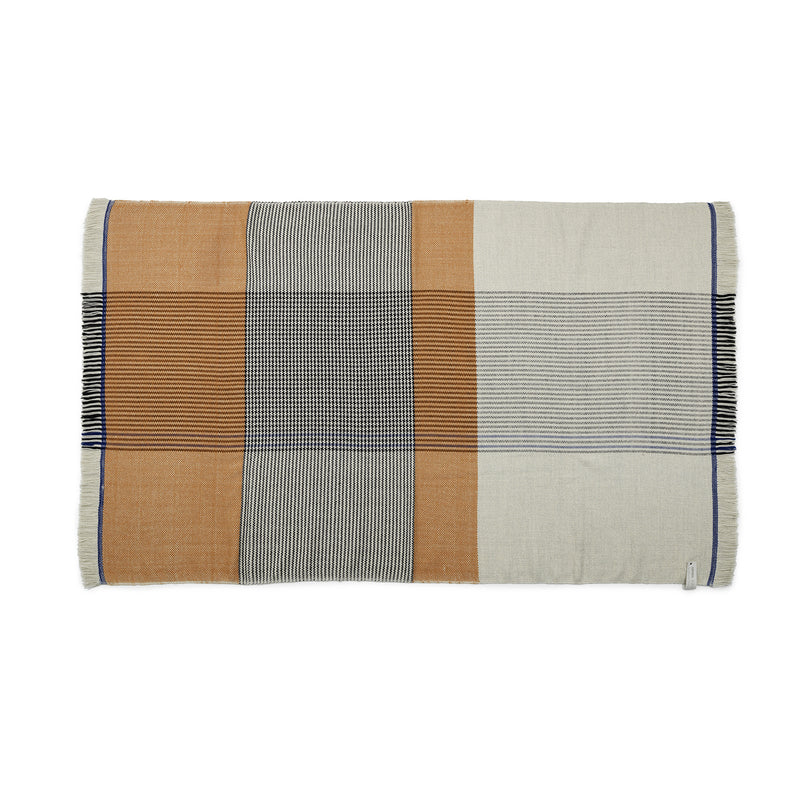 The Ames Ruana Throw is a rectangular woven blanket that uses traditional techniques and earthy tones. It features vertical and horizontal stripes in beige, brown, black, cream patterns, fringed edges like Ruana throws, and a small fabric tag on one side.
