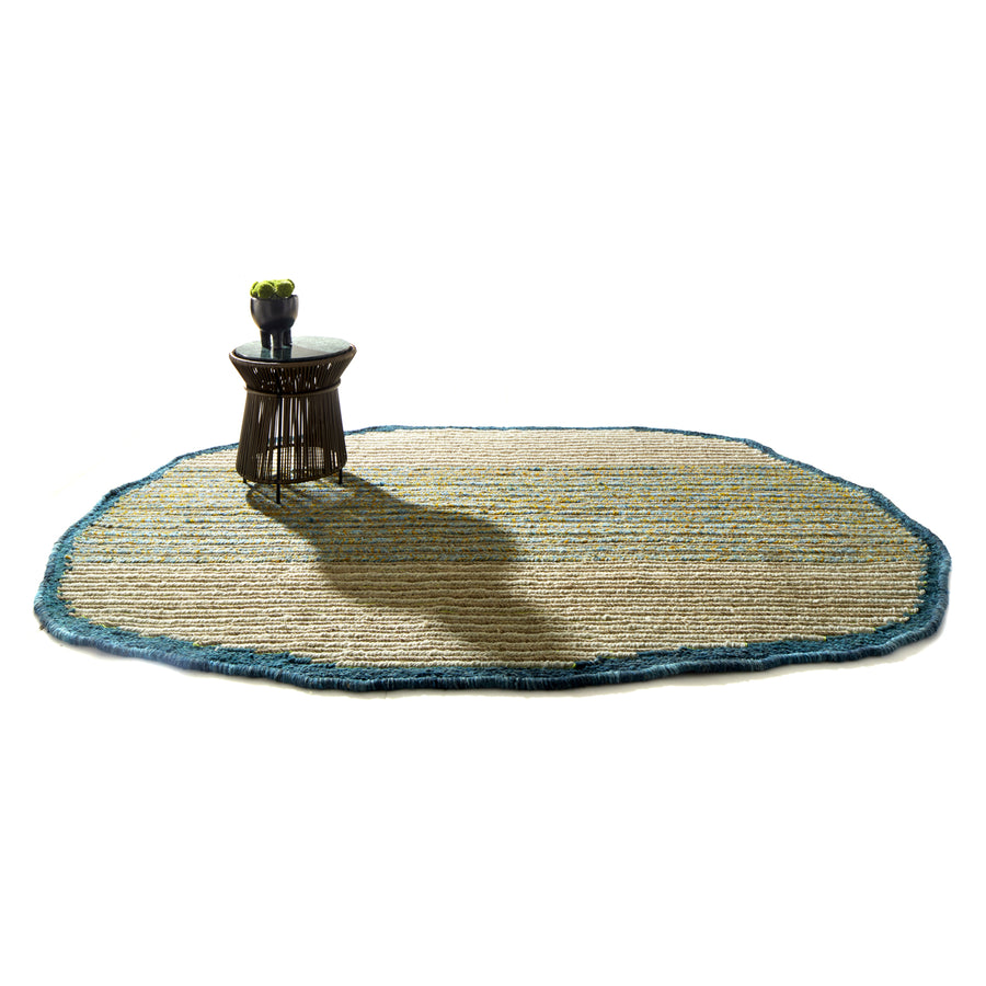 A small round table with a bowl of green apples sits on an irregularly shaped Ames Uilas Rug by Ames, featuring blue edges, casting a shadow on a white background.