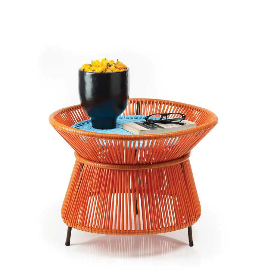 The Caribe Basket Table from Ames features a unique vase-like shape with thin black legs. This orange, woven wicker table from the Caribe Collection highlights traditional Momposino weaving and holds a black pot with yellow peppers, a closed book, and black sunglasses on a blue mat.