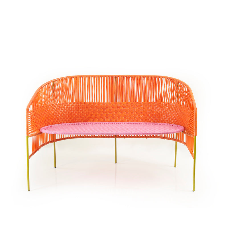 The Caribe Sofa by Ames features a modern orange woven design using traditional momposino techniques. It includes a pink cushion and rounded back, supported by four slender yellow legs. Ideal as Caribbean coastal outdoor furniture, it is beautifully set against a plain white backdrop.
