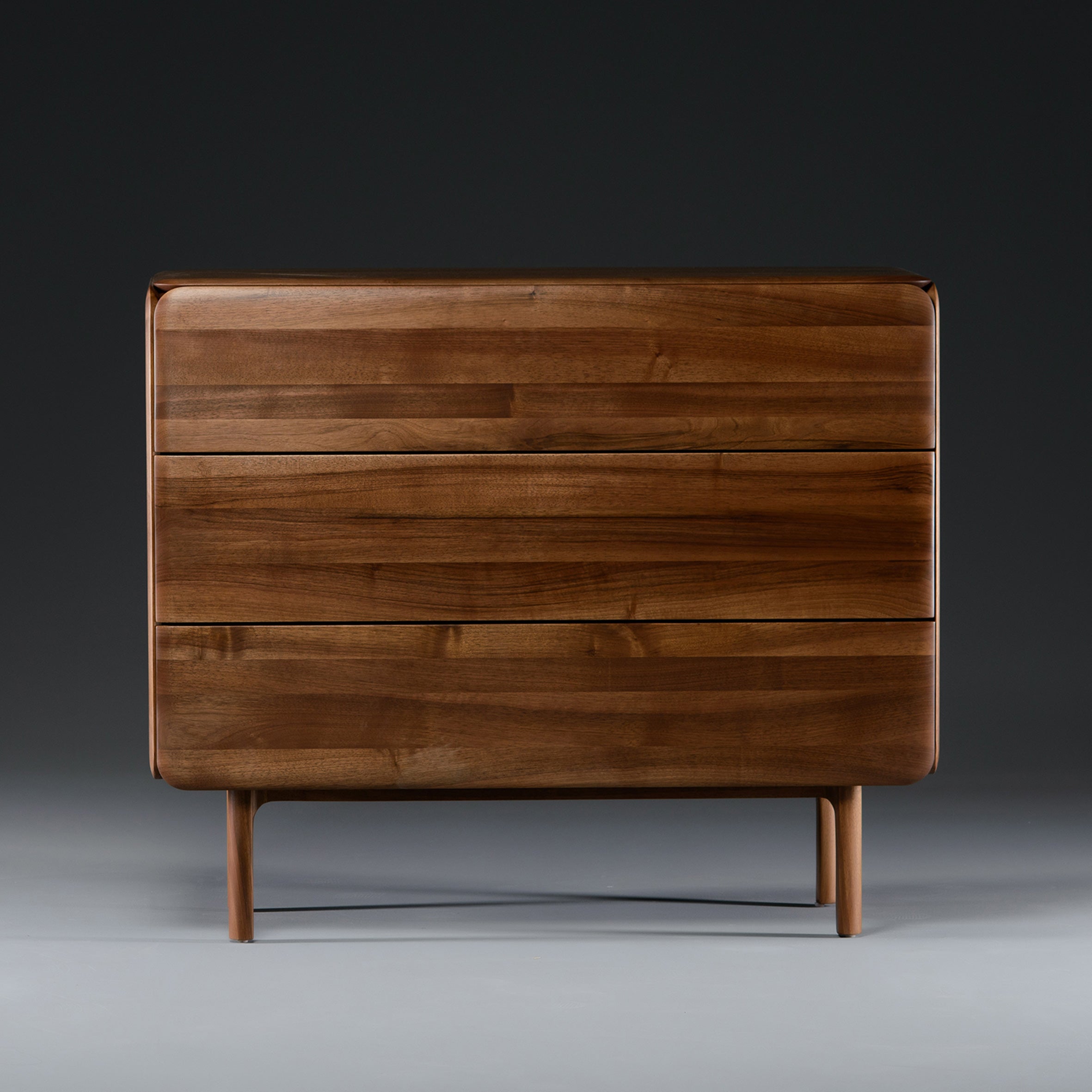 The CLOUD High board by Artisan is a modern, solid wood dresser with a dark finish made from eco-friendly materials. It has three large drawers, a minimalist design with rounded edges, and slender legs against a dark grey background.