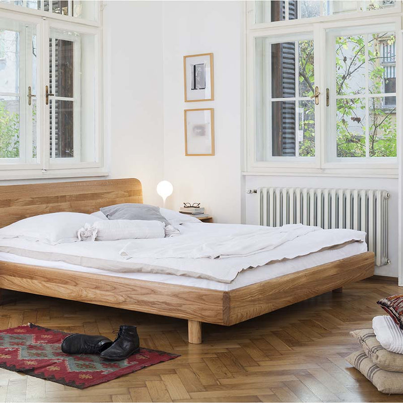 The minimalist bedroom features the Artisan FIN Bed with white bedding, complemented by a small bedside lamp and framed artwork. Large windows highlight greenery outside, while a patterned rug and a pair of black shoes complete the scene.