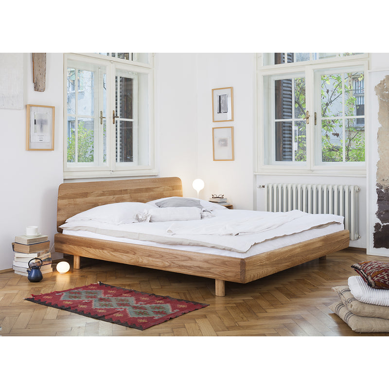 The minimalist bedroom features the Artisan FIN Bed and two small tables with lamps. Large windows light up the white walls, framed pictures, and a red patterned rug on the wooden floor. Books and a tea set by the bed complement the eco-friendly materials.
