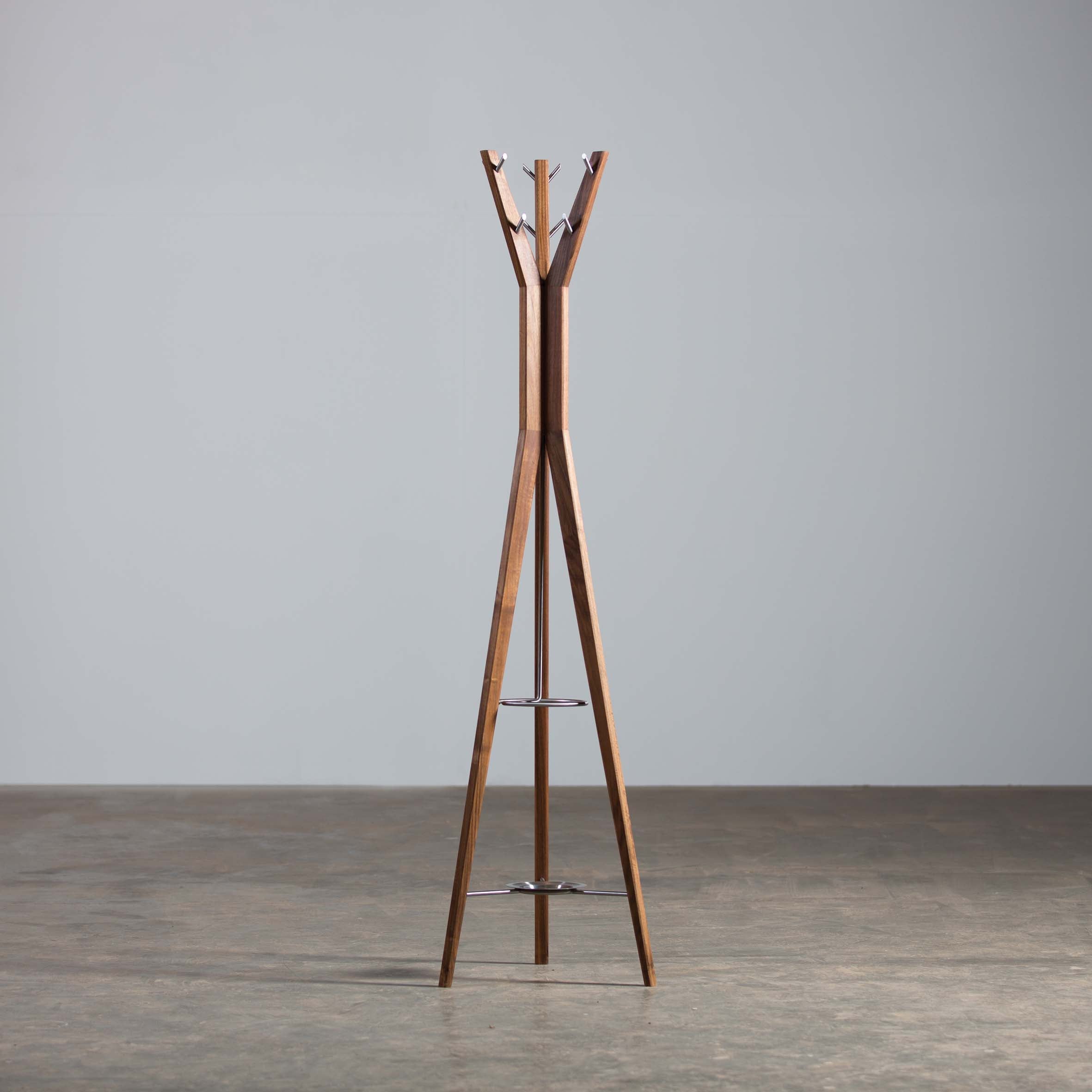 The Artisan HANNY Coat Stand, featuring a tall, modern design with a tripod base, stands on a concrete floor against a light gray wall. Crafted from solid wood with metal hooks and a sleek finish, this minimalist piece is made using environmentally-friendly materials.