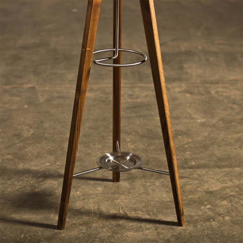 The HANNY Coat Stand by Artisan is an elegant, eco-friendly piece made from solid wood. It features three tall wooden legs with metal supports, a bottom metal dish, and an upper ring for holding objects. Its beautifully showcased against a textured concrete surface.