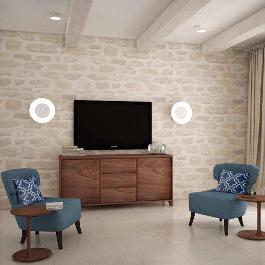 A modern living room with a stone accent wall features a flat-screen TV, two blue armchairs with patterned pillows, and the Artisan HANNY Sideboard crafted from solid wood. Circular wall lights flank the TV. Environmentally-friendly materials in neutral tones dominate the space.