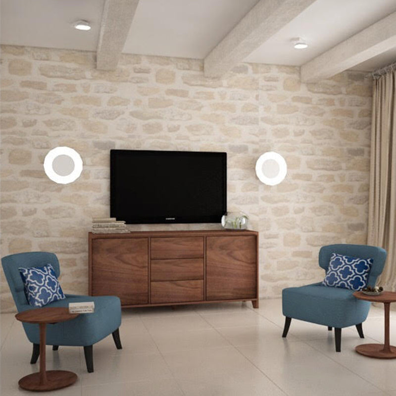 A modern living room with a stone accent wall features a flat-screen TV, two blue armchairs with patterned pillows, and the Artisan HANNY Sideboard crafted from solid wood. Circular wall lights flank the TV. Environmentally-friendly materials in neutral tones dominate the space.