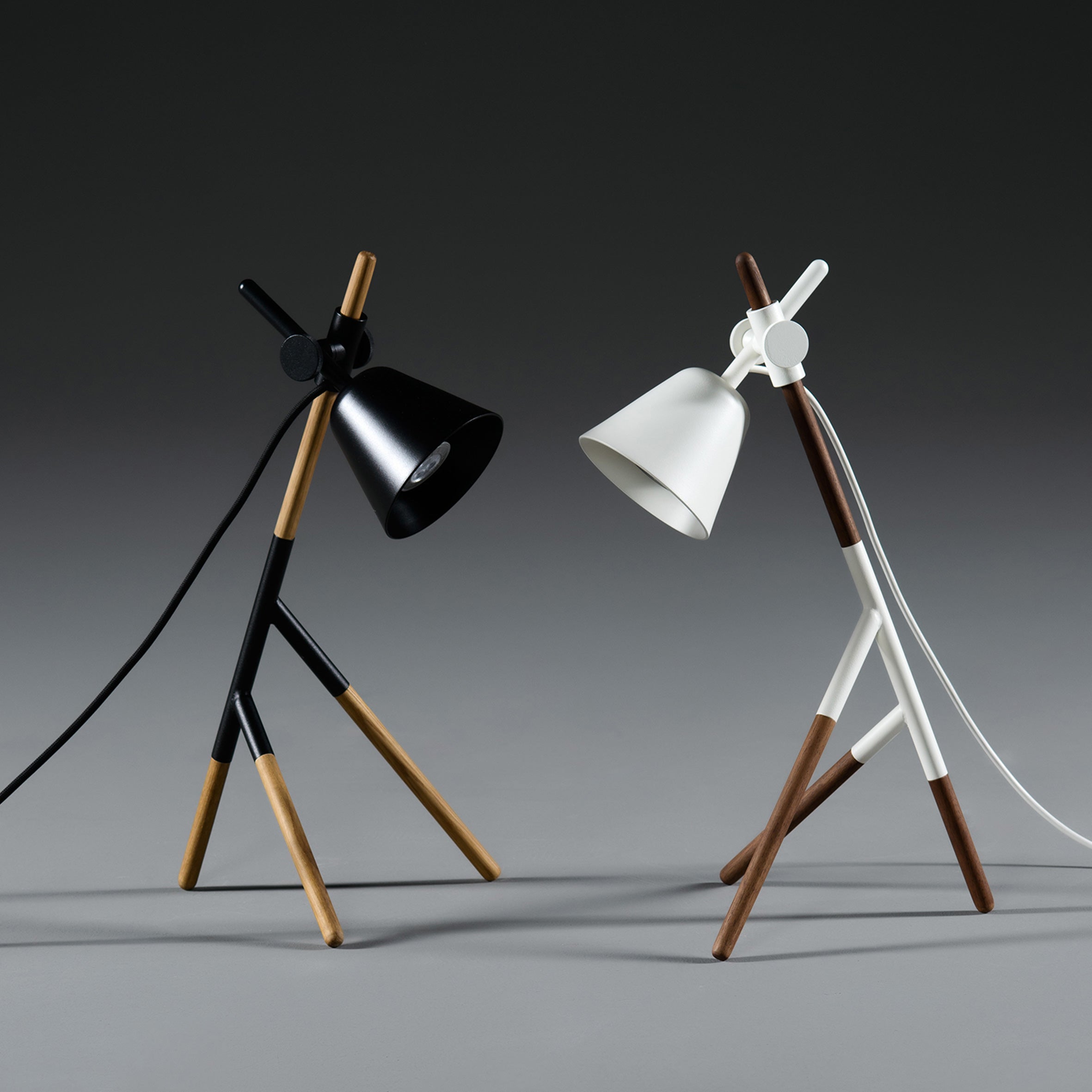 The Artisan INSERT Table Lamps feature a minimalist design with eco-friendly solid wood legs in black and white, each with angled shades. Set against a neutral gray backdrop, the stylish tripod lamps seem to lean toward each other.