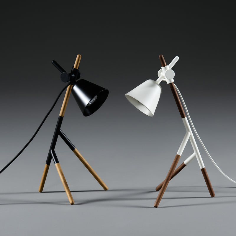 The Artisan INSERT Table Lamps feature a minimalist design with eco-friendly solid wood legs in black and white, each with angled shades. Set against a neutral gray backdrop, the stylish tripod lamps seem to lean toward each other.