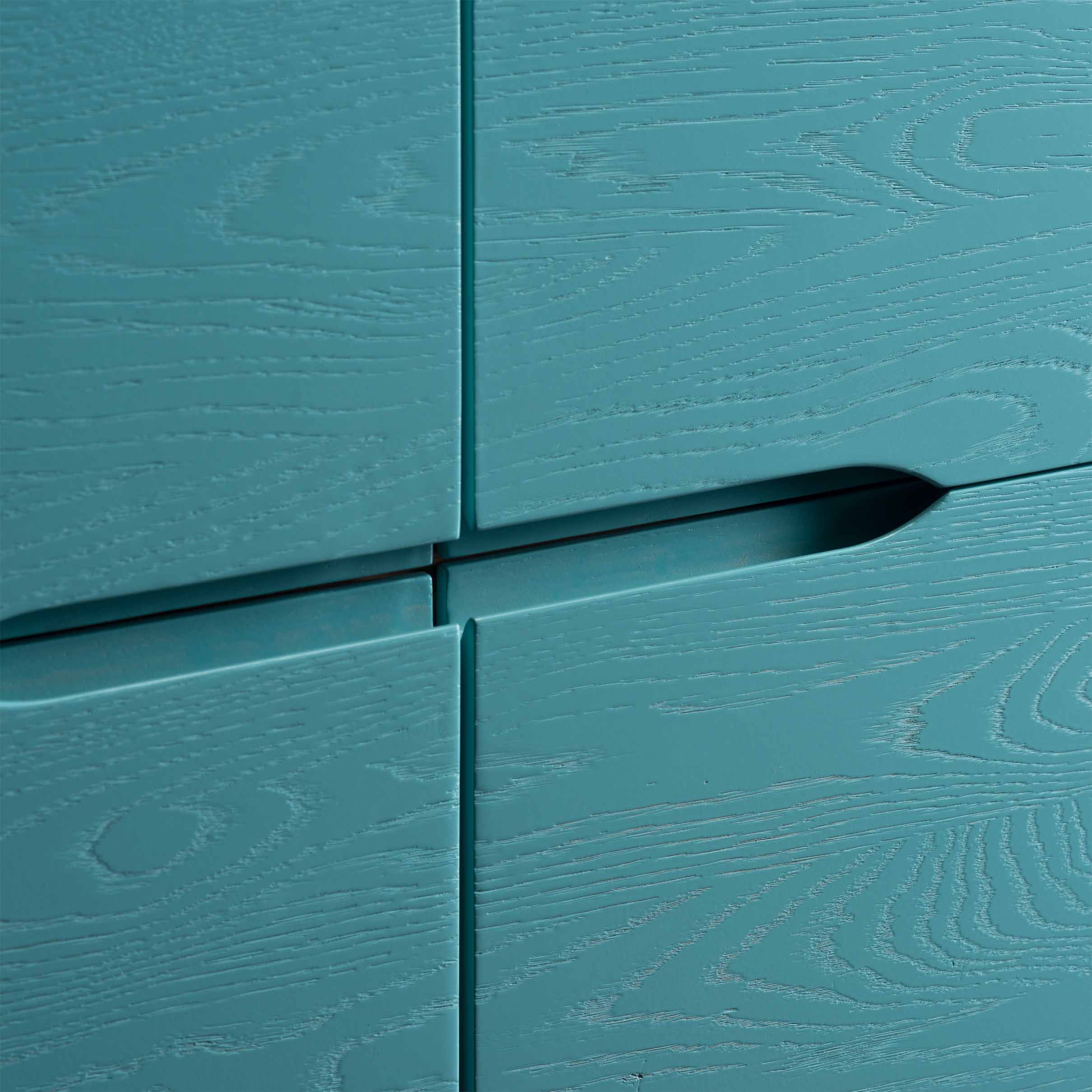 Close-up of the Artisan INVITO Highboard, a teal wooden cabinet with visible grain patterns and integrated handles. The image highlights the join between two drawers, showcasing its textured surface and sleek, modern design crafted from environmentally-friendly solid wood.