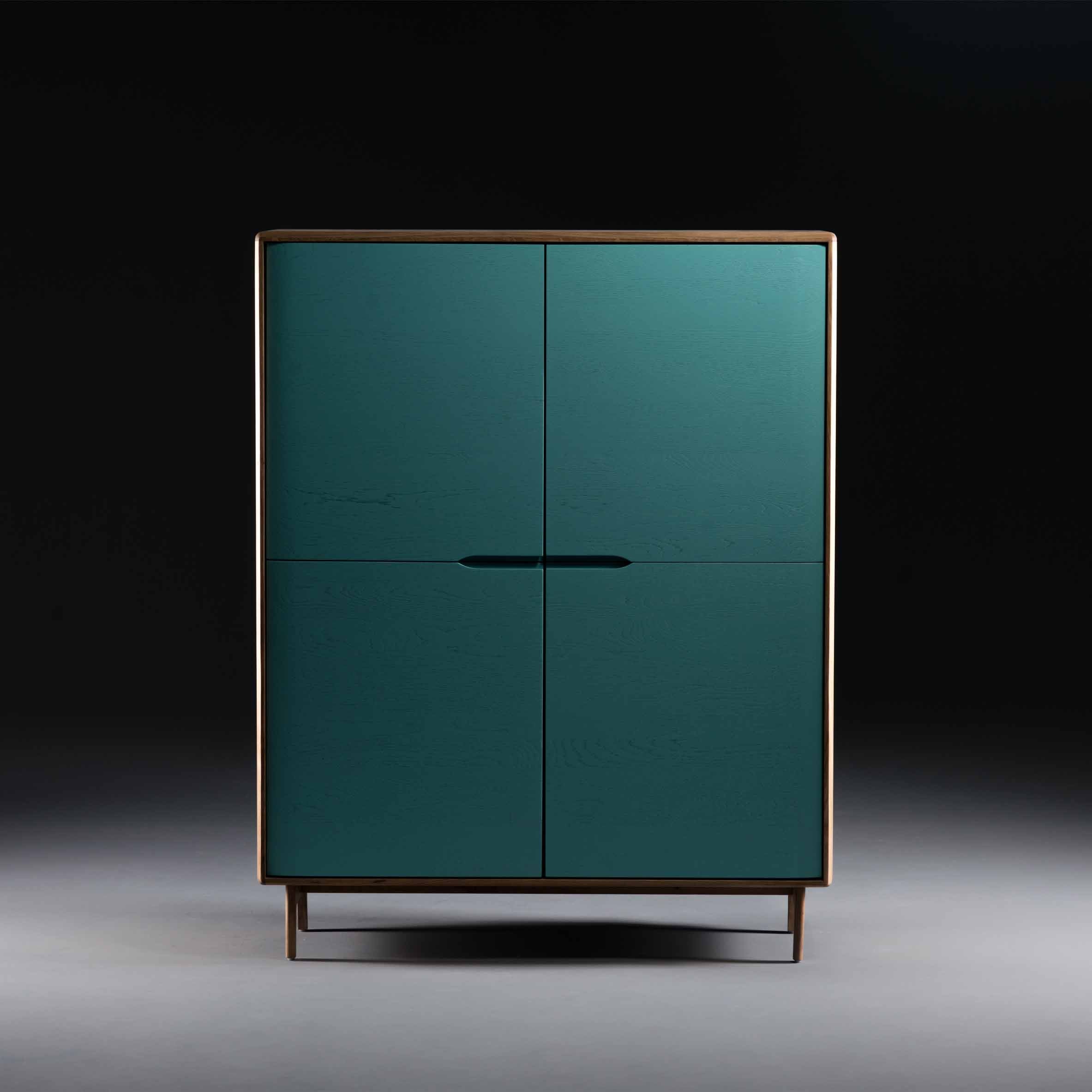 The INVITO Highboard by Artisan is a sleek, modern cabinet with a minimalist design, featuring a solid wood frame and teal doors. Crafted from eco-friendly materials, it stands on slim legs that cast subtle shadows against a dark gradient backdrop.