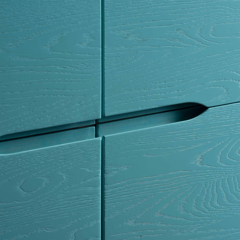 Close-up of the Artisan INVITO Highboard, a teal wooden cabinet with visible grain patterns and integrated handles. The image highlights the join between two drawers, showcasing its textured surface and sleek, modern design crafted from environmentally-friendly solid wood.