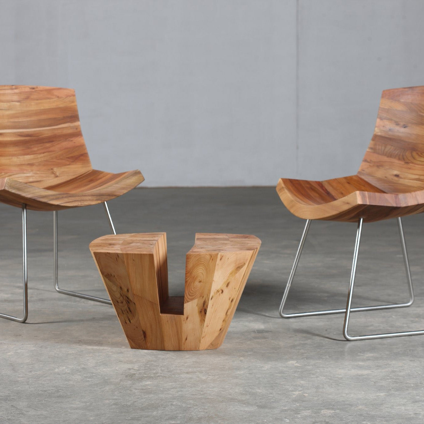 Two modern wooden chairs with curved seats and metal legs flank the KART Coffee Table by Artisan, crafted from solid wood, on a concrete floor. The minimalist setting, enhanced by a light gray wall, exudes an environmentally-friendly vibe with natural oils.