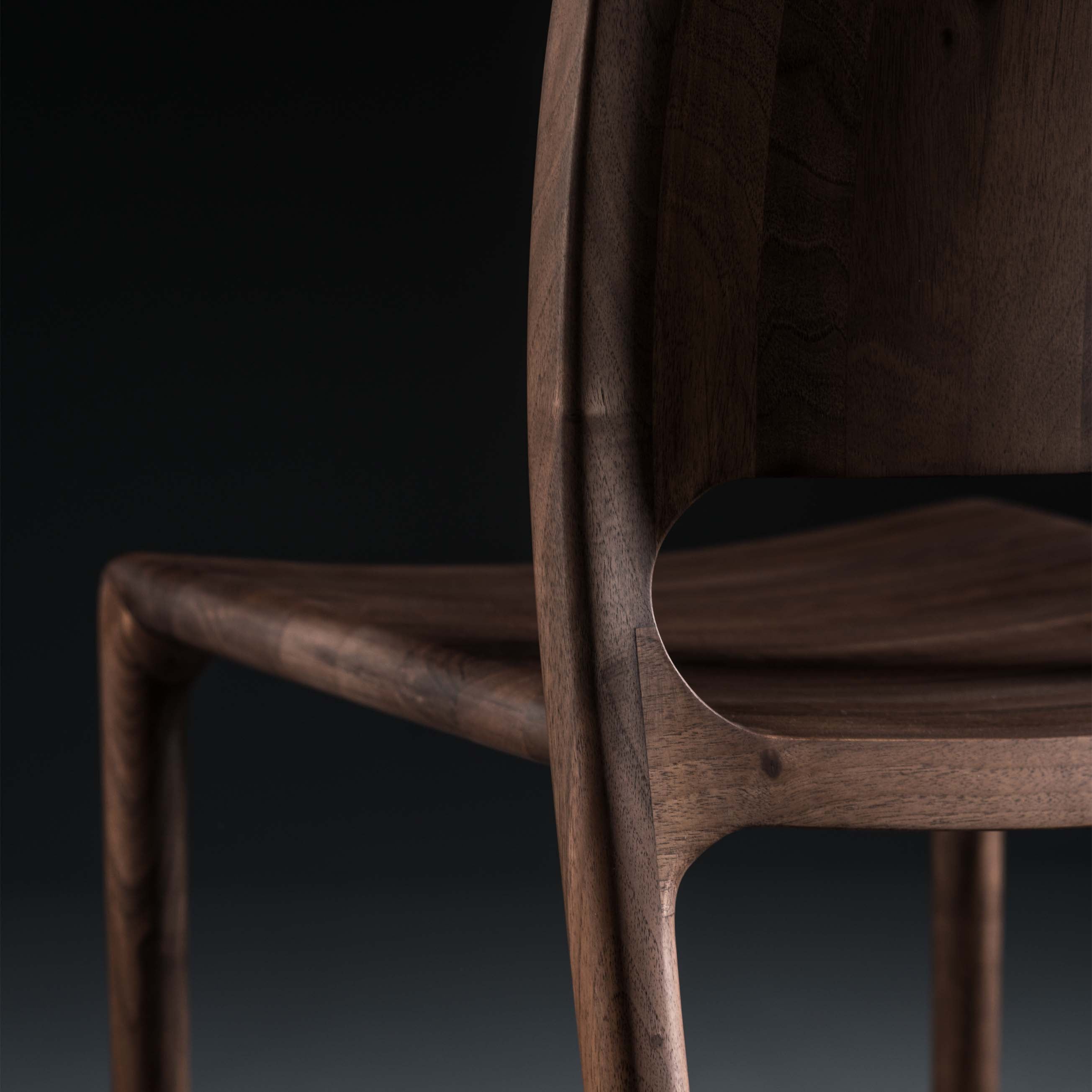 A close-up of the LATUS Chair by Artisan features its smooth, curved backrest and seat. Crafted from solid wood, the rich brown grain and natural oils stand out against a dark background, highlighting its beauty and eco-friendliness.