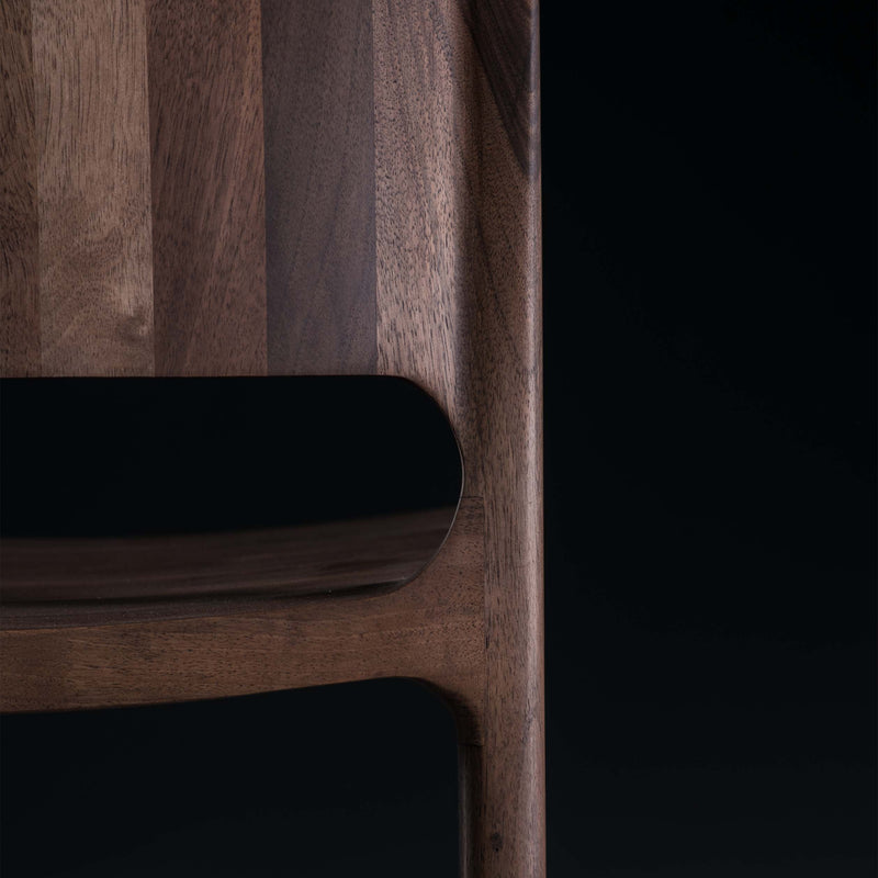 Close-up of the LATUS Chair by Artisan, featuring a portion of its curved backrest and legs. Crafted from solid wood, its rich brown grain and polished finish are enhanced by natural oils, set against a dark background.