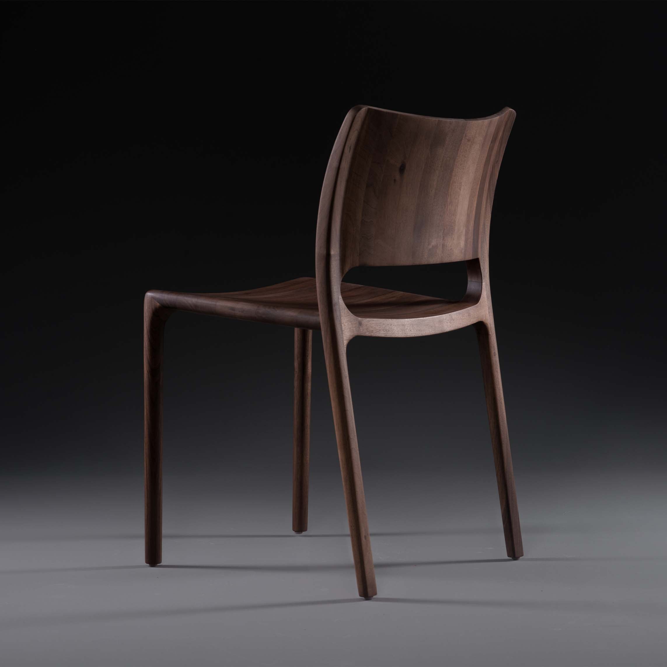 The LATUS Chair by Artisan is a sleek, modern piece crafted from solid wood featuring a curved backrest and minimalist design. Shown at an angle against a dark backdrop, its rich grain shines with natural oils, enhancing its elegant and eco-friendly aesthetic.