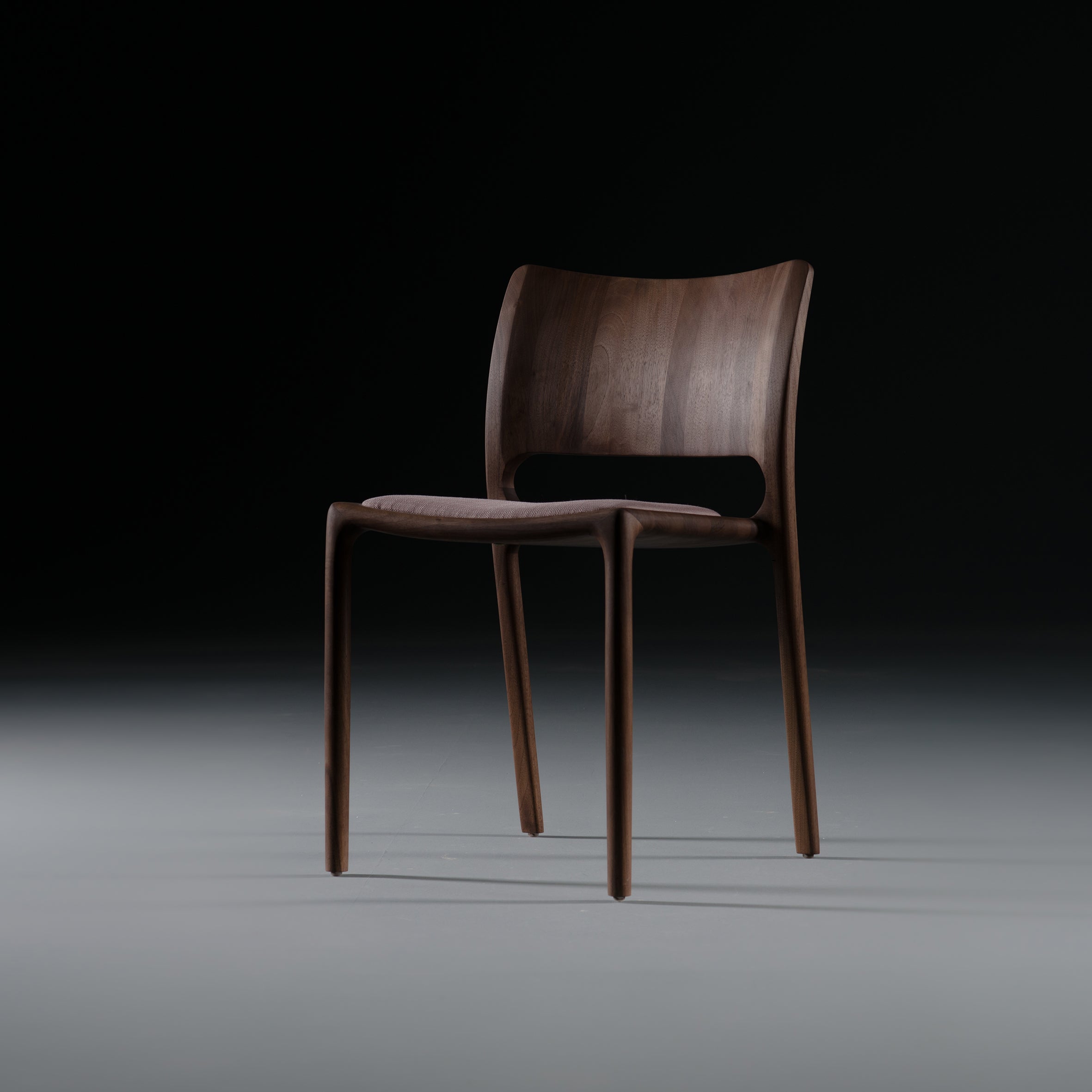 The LATUS Chair by Artisan, crafted from solid wood, features a minimalist design with a curved backrest. Set on a gray floor against a dark background, its smooth finish is accentuated by natural oils and elegant form highlighted by subtle lighting.