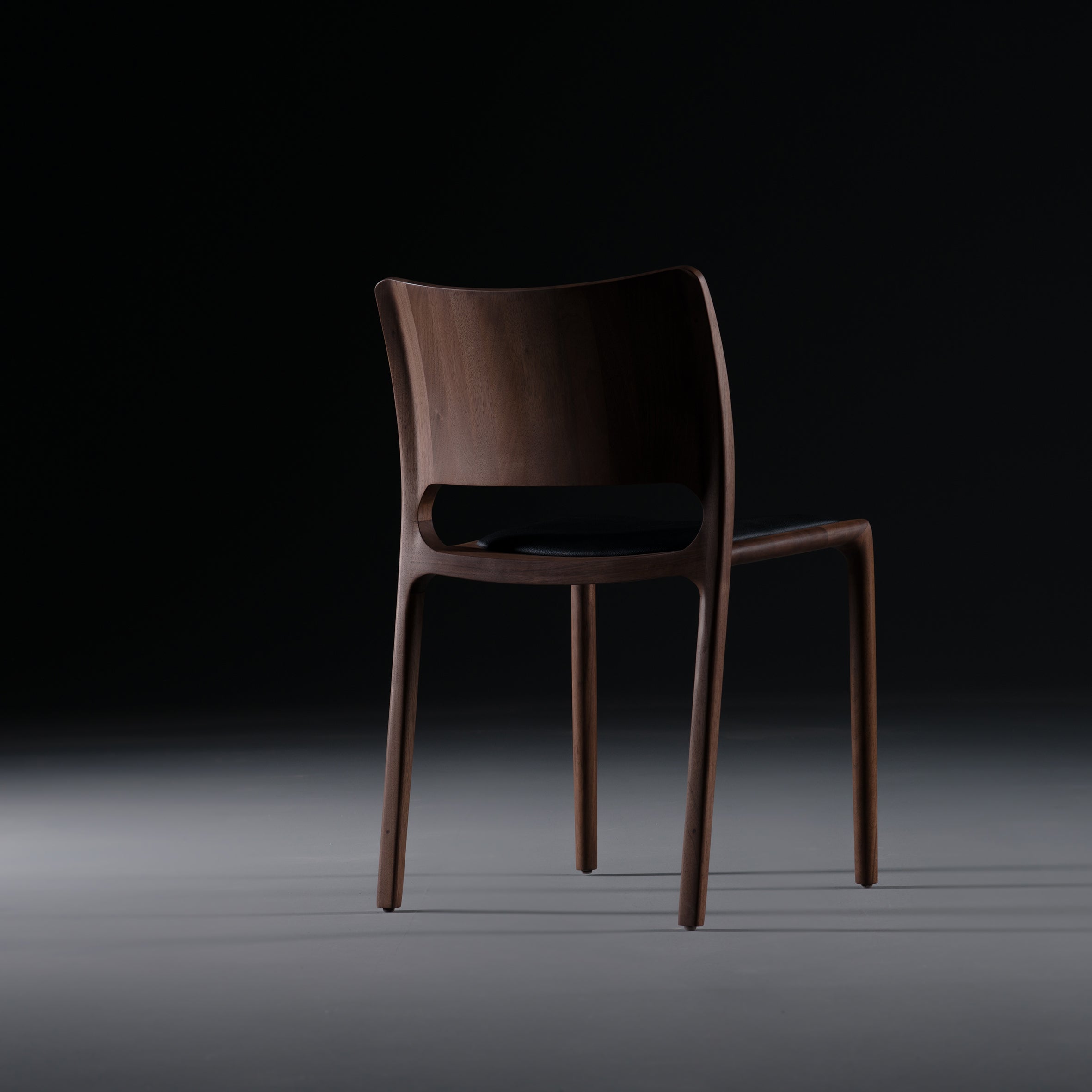 The LATUS Chair by Artisan, crafted from solid wood and enhanced with natural oils, showcases a sleek modern design with a curved backrest and elegant lines. Its positioned on a dimly lit surface against a dark background.