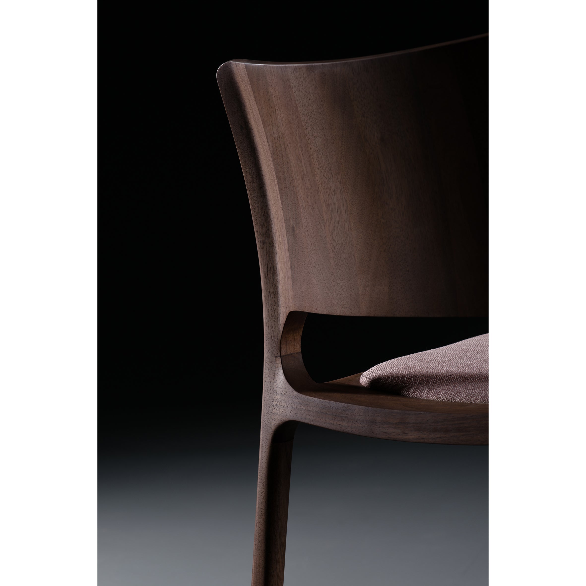 Close-up of the Artisan LATUS Chair, made from solid wood with natural oils, set against a dark background. It showcases smooth curves, a curved backrest, and a soft, muted fabric seat. The lighting highlights its wood grain and design details.